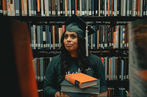 Student sitting next to bookshelf wearing graduation cap